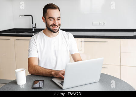 Heureux bel homme freelancer assis à la table de cuisine, working on laptop computer Banque D'Images