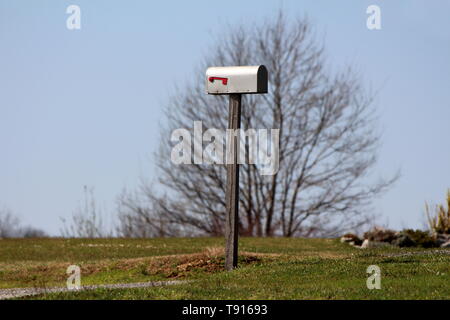 Boîte aux lettres en métal gris monté sur poteau en bois en haut de la petite colline entourée d'herbe et de grand arbre sur ciel bleu clair en arrière-plan Banque D'Images