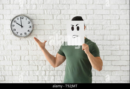 Jeune homme se cachant derrière feuille de papier, appelée émoticône contre mur en brique blanche avec horloge Banque D'Images