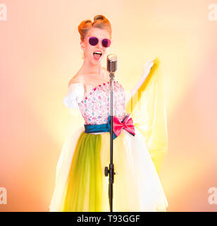 Happy girl singer en lunettes roses sourit, s'amuser, chante en retro microphone, montre les points à la main doigt dans l'appareil photo. Adolescent élégant musicien dans Banque D'Images