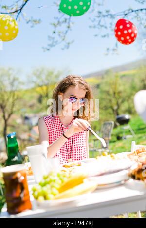 De teen girl in red chemise à carreaux assis par table sur anniversaire garden party de manger avec fourchette en plastique Banque D'Images