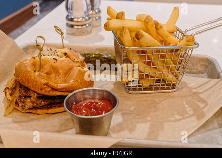 Entrecôte grillée et oignons frits sur un rouleau rond avec un petit panier de frites, du ketchup et un cornichon sur un plateau de service en métal avec un papier parchemin Banque D'Images