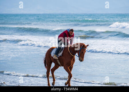 Ayia Eirini, Chypre - 24 mars, 2019 : Man riding marron sur un cheval au galop sur la plage d'Ayia Erini à Chypre contre une mer rugueuse Banque D'Images