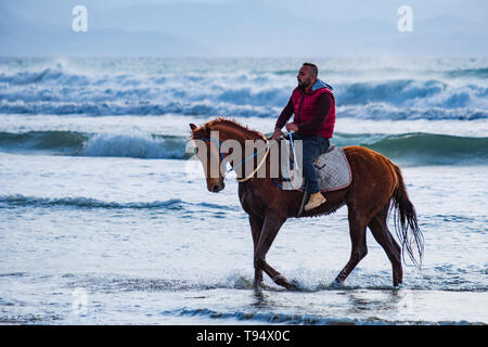 Ayia Eirini, Chypre - 24 mars, 2019 : Man riding marron sur un cheval au galop dans les eaux de mer de la plage de Ayia Erini à Chypre contre une mer rugueuse Banque D'Images