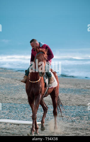 Ayia Eirini, Chypre - 24 mars, 2019 : Man riding marron sur un cheval au galop sur la plage d'Ayia Erini à Chypre contre une mer rugueuse Banque D'Images