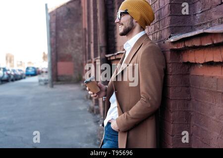 Beau jeune homme dans des vêtements décontractés holding disposable cup et souriant tout en marchant à travers la ville. Banque D'Images