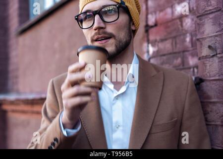 Beau jeune homme dans des vêtements décontractés holding disposable cup et souriant tout en marchant à travers la ville. Banque D'Images