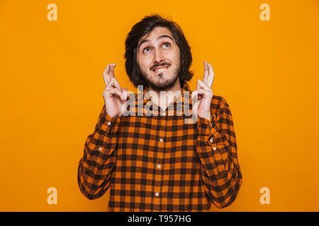 Portrait de jeune homme priant chemise à carreaux en gardant les doigts croisés en se tenant debout sur fond jaune isolé en studio Banque D'Images