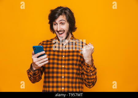 Photo de young man wearing plaid shirt rire et à l'aide du téléphone cellulaire isolés sur fond jaune en studio Banque D'Images