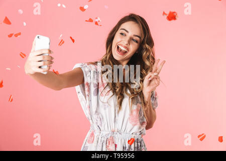 Image de femme joyeuse 20s robe porter et souriant en tenant sous les confettis selfies, isolé sur fond rose Banque D'Images