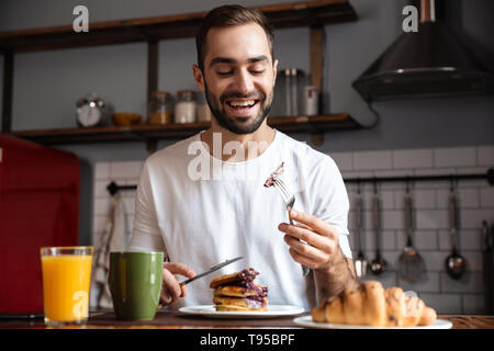 Image de brunette guy 30s assis à table et de manger des aliments pendant le petit-déjeuner à appartement moderne Banque D'Images