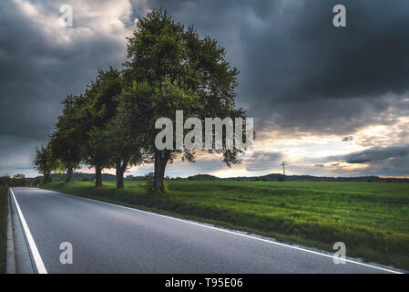 La rue vide, entourée d'arbres, sous un ciel d'orage, avec le soleil perçant les nuages, traversant des champs verts, près de Schwabisch Hall, en Allemagne. Banque D'Images