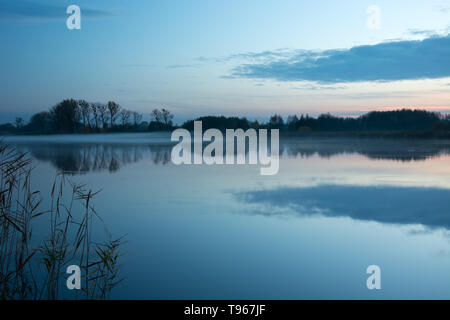 Le brouillard sur le lac et arbres sur la rive se reflétant dans l'eau après le coucher du soleil Banque D'Images