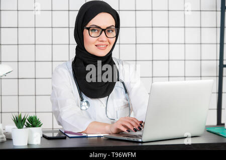 Femme scientifique voilée assise sur son bureau avec les doigts sur un clavier de son ordinateur portable, en souriant. Banque D'Images