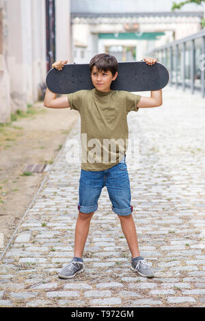 Jeune garçon joyeux dans la rue tout en tenant une planche à roulettes tout en regardant loin dans un jour lumineux Banque D'Images
