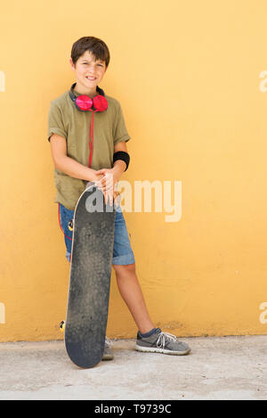 Young boy leaning on mur jaune avec des écouteurs sur le cou, tenant une planche à roulettes tout en regardant loin dans un jour lumineux Banque D'Images