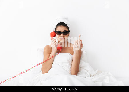 Jeune femme en colère après la douche assis au lit enveloppé dans une couverture, le port de lunettes, de parler sur un téléphone fixe Banque D'Images