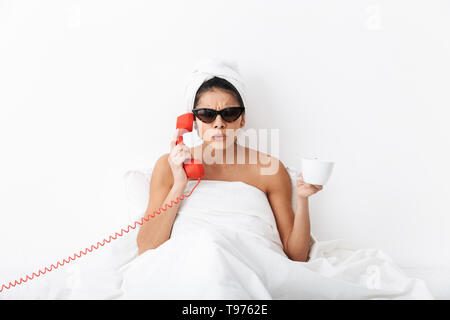 Jeune femme en colère après la douche assis au lit enveloppé dans une couverture, le port de lunettes, de parler sur un téléphone fixe, tenant une tasse Banque D'Images