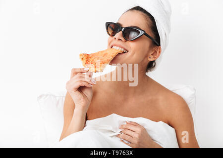 Cheerful young woman sitting in bed après douche enveloppé dans une couverture, le port de lunettes de soleil, manger tranche de pizza Banque D'Images