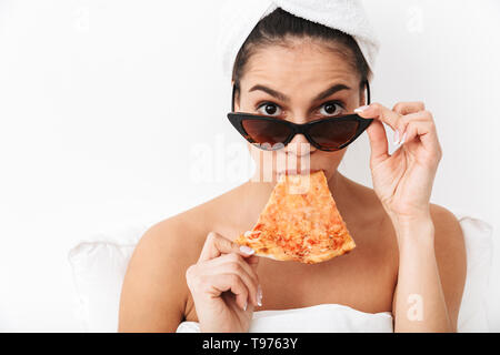 Cheerful young woman sitting in bed après douche enveloppé dans une couverture, le port de lunettes de soleil, manger tranche de pizza Banque D'Images