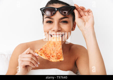 Cheerful young woman sitting in bed après douche enveloppé dans une couverture, le port de lunettes de soleil, manger tranche de pizza Banque D'Images