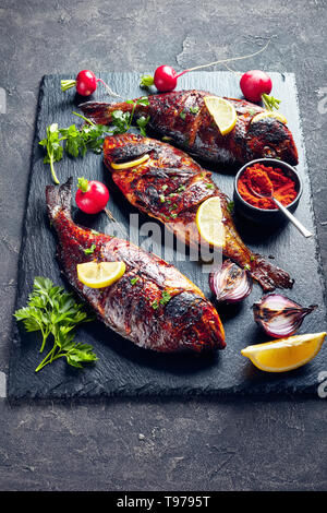 Trois délicieux grillés fraîchement dorado poisson servi sur un plateau en ardoise noire avec des radis, oignons rouges grillés et des rondelles de citron sur une table en béton, vertical Banque D'Images