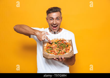 Image de l'homme optimistes en 30s blanc T-shirt holding et de manger une pizza tout en isolés sur fond jaune Banque D'Images