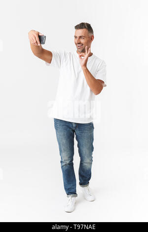 Longueur totale de l'image 30s homme unshaved portant des T-shirt et jeans prendre photo selfies sur téléphone mobile tout en tenant dans la main isolated over white b Banque D'Images