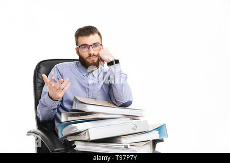 Jeune homme élégant barbu ennuyé à lunettes assis sur un fauteuil avec les dossiers et regardant la caméra sur fond gris Banque D'Images