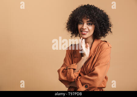 Jolie femme africaine joyeuse de toucher son menton et regardant la caméra sur fond brun Banque D'Images