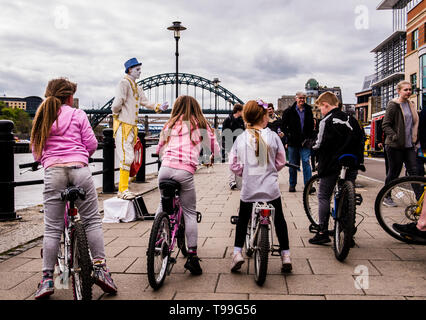 Vue arrière du groupe d'enfants faire du vélo aux côtés de Quay, artiste de rue en face d'eux, Quayside, Newcastle upon Tyne, England, UK Banque D'Images