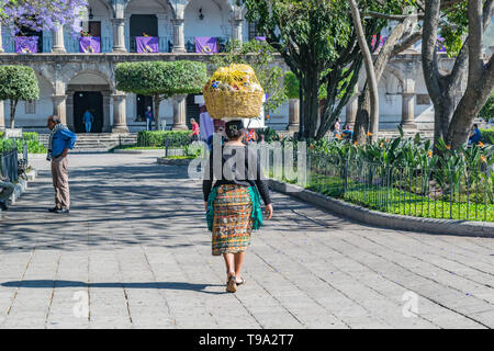 Femme dans une cuisine guatémaltèque les vêtements, marcher dans Central Park avec un panier sur la tête et un bâtiment historique, à Antigua, Guatemala Banque D'Images