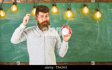 Homme avec barbe et moustache sur visage oublieux des démangeaisons tête. Hipster barbu est titulaire d'réveil, tableau sur toile de fond, l'espace de copie. Concept de discipline. Enseignant dans les lunettes est titulaire d'un réveil et un stylo. Banque D'Images