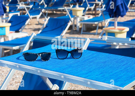 Lunettes de soleil hommes et femmes sur une chaise longue bleu vif avec une vue de flou artistique d'une rangée de chaises longues, parasols et plage de galets Banque D'Images