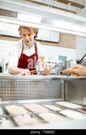 Jeune homme en tablier rouge vend de la crème glacée, montrant différentes saveurs dans la pâtisserie Banque D'Images