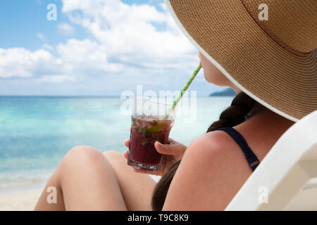 Femme assise sur une chaise Wearing Hat boire le jus avec de la paille à la plage Banque D'Images