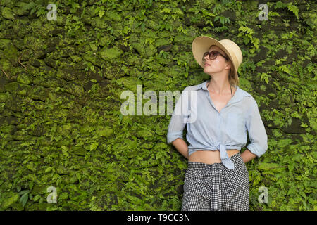 Portrait d'une belle femme traveler. Smiling young woman in summer hat portant des lunettes, debout devant des plantes tropicales luxuriantes de verdure mur Banque D'Images