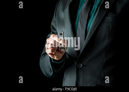 Close-up of man in black incognito, holding vintage clé avec deux doigts, isolés sur fond noir. Banque D'Images