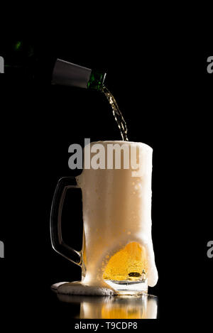 Verre à bière débordante avec un liquide est versé dans une bouteille au-dessus de façon à ce que la mousse déborde sur un compteur de réflexion sombre sur un fond noir. Banque D'Images