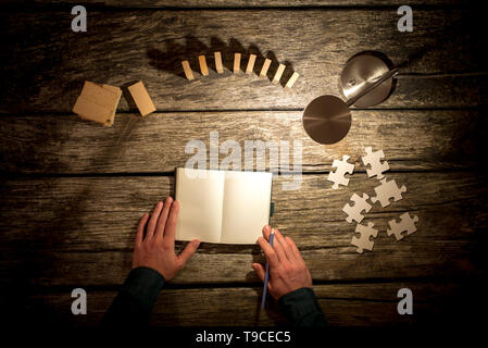 Vue du dessus de l'homme sur les mains pour prendre des notes en blanc ordinateur portable et divers piquets, dominos et pièces de casse-tête placé autour de lui sur l'étude en bureau avec ta Banque D'Images