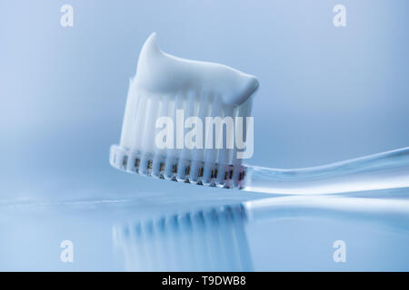 Brosse à dents en plastique transparent avec du dentifrice blanc sur un fond blanc bleu avec la réflexion sur le verre. Banque D'Images