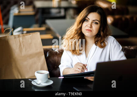 Une jeune femme, sympathique, pas une mince-dirigé musculation, est assis dans un café confortable, fonctionne sur un ordinateur et navigue sur des documents. Banque D'Images