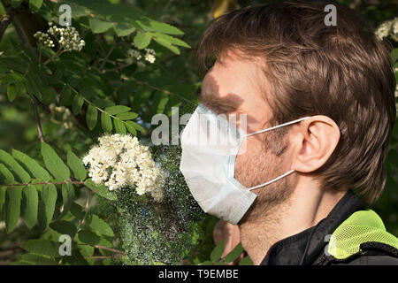 Homme séduisant avec masque médical sur son visage, l'ombre dans ses yeux contre nuage de pollen d'un arbre en fleurs Banque D'Images