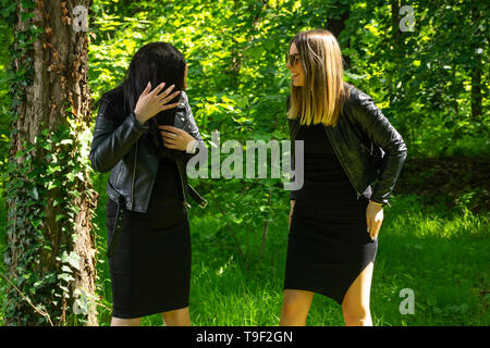 Deux heureux filles rient avec dents sourire au printemps journée ensoleillée dans la forêt. Une fille changer les cheveux. Filles portent une robe noire et veste en cuir. Close up Banque D'Images