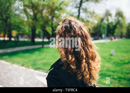 Close-up of young woman avec les cheveux bouclés walking in park sur jour de printemps ensoleillé, vue de dos. Banque D'Images