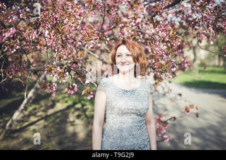 Happy middle aged woman arménienne dans une élégante robe sous l'arbre sakura en fleurs. Souriant, à côté. Banque D'Images