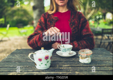 Une jeune femme est de boire du thé à l'extérieur dans un parc et est en remuant sa tasse Banque D'Images