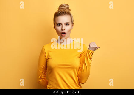 Fille émotionnelle étonné avec bouche ouverte avec des points de côté du pouce, a surpris l'expression du visage, isolé sur fond jaune.voir ici Banque D'Images