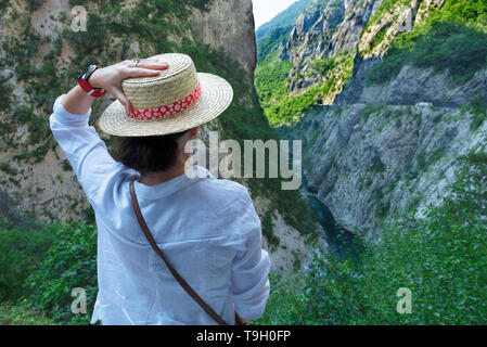 Une jeune femme en blouse blanche et chapeau de paille regarde la rivière de montagne qui s'écoule entre la gorge de pierre. Banque D'Images