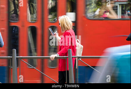 Belgrade, Serbie - Avril 22, 2019 : Young blonde woman looking at son téléphone portable lors de l'attente à l'arrêt de bus sur la rue de la ville sur une journée de printemps ensoleillée avec Banque D'Images